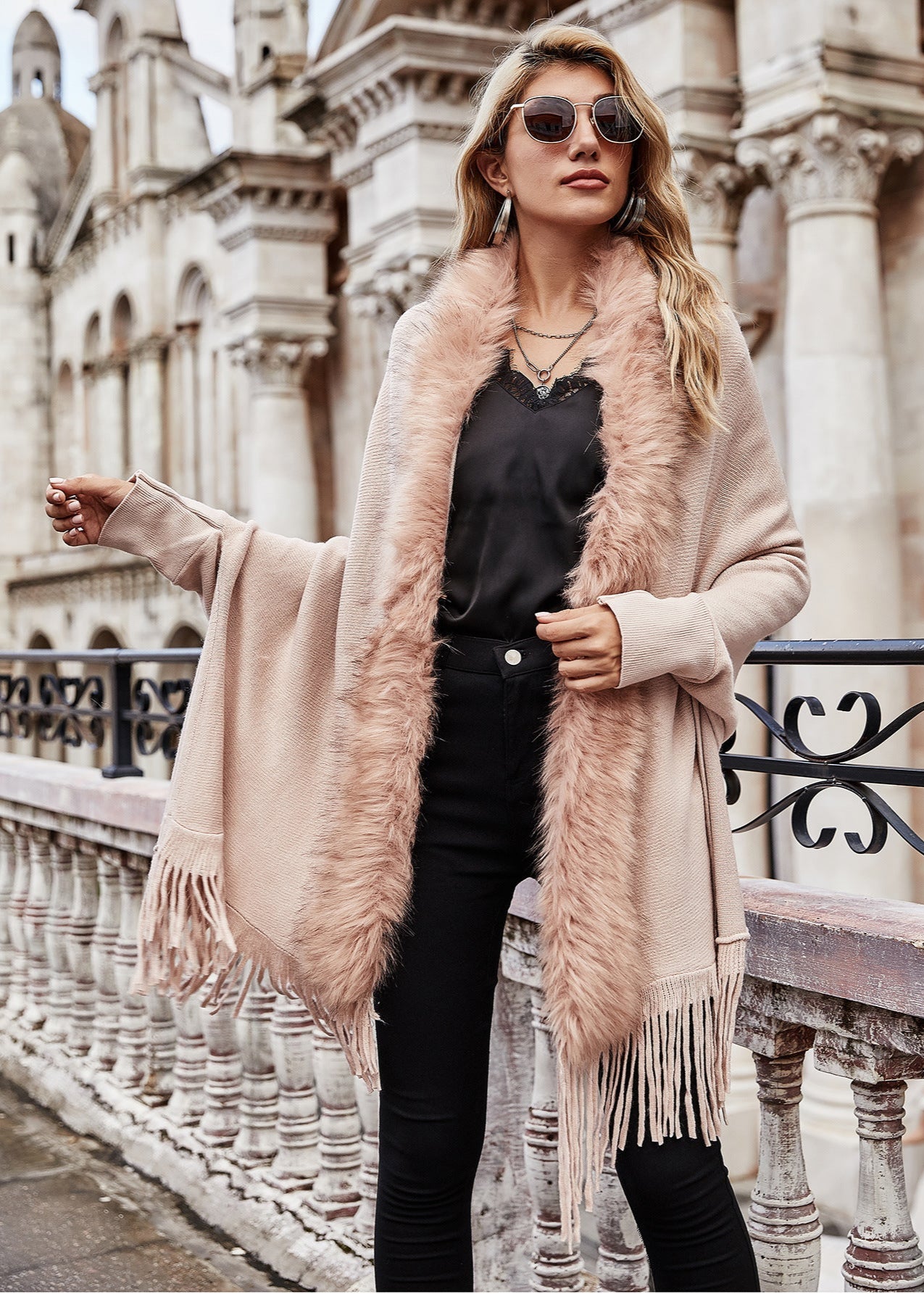 Woman wearing a pink fur-trimmed coat and sunglasses, standing in front of an architectural building.