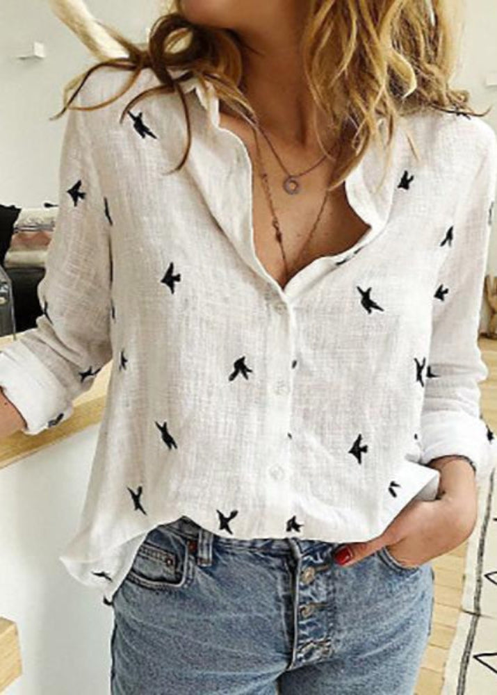 Woman wearing a white blouse with black patterns and blue jeans indoors.