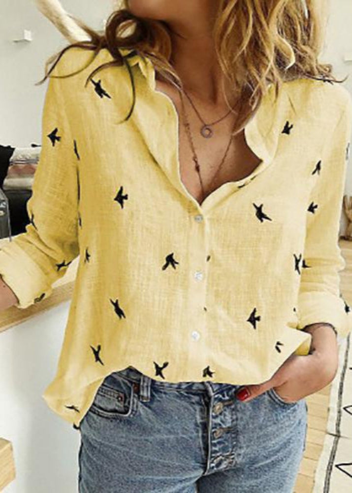 Woman wearing a yellow blouse with black patterns and blue jeans indoors.