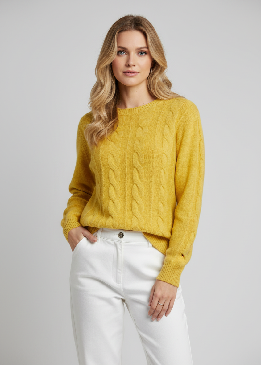 Woman wearing a mustard yellow cable knit sweater and white pants on a plain background