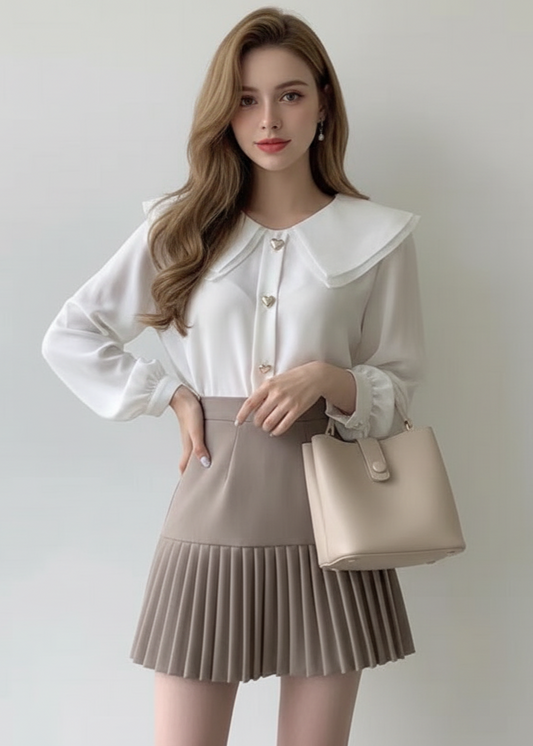 Woman wearing a white blouse with a large collar and a beige pleated skirt, holding a beige handbag against a plain background.