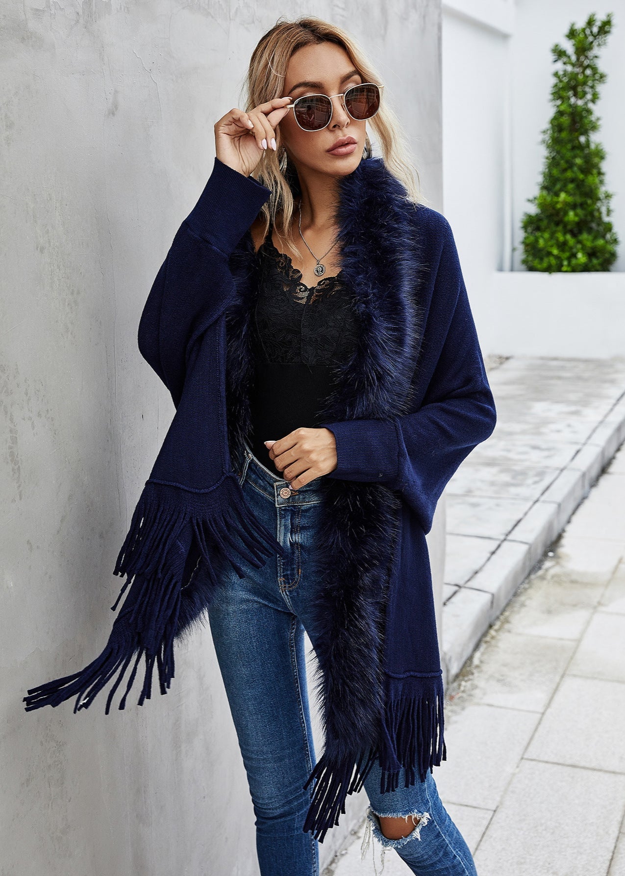 Woman wearing a navy blue fringed jacket with sunglasses, standing against a light gray wall.